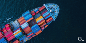 Aerial view of a large cargo ship transporting multicolored shipping containers across the ocean. The vessel, loaded with red, blue, yellow, and orange containers, is sailing through deep blue waters, symbolizing global trade, maritime logistics, and international shipping.