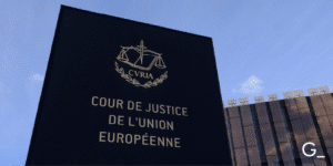 The image shows the exterior of the Court of Justice of the European Union (Cour de Justice de l'Union Européenne). The sign features the emblem of the European Court of Justice with the scales of justice symbol and the abbreviation CVRIA. The building appears modern with a glass facade and is set against a clear blue sky.