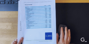A person holds a financial report; on the left is a laptop keyboard, on the right a mouse on a black mat, with a G-BRIS logo in the page’s right corner